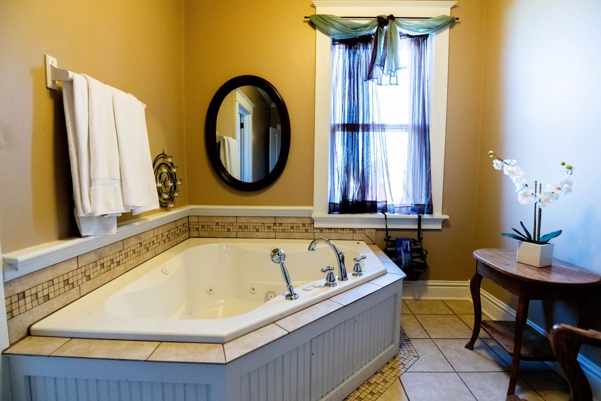 This elegant bathroom features a corner jetted tub, a round mirror, and a window with sheer dark blue drapes, complemented by light brown walls.