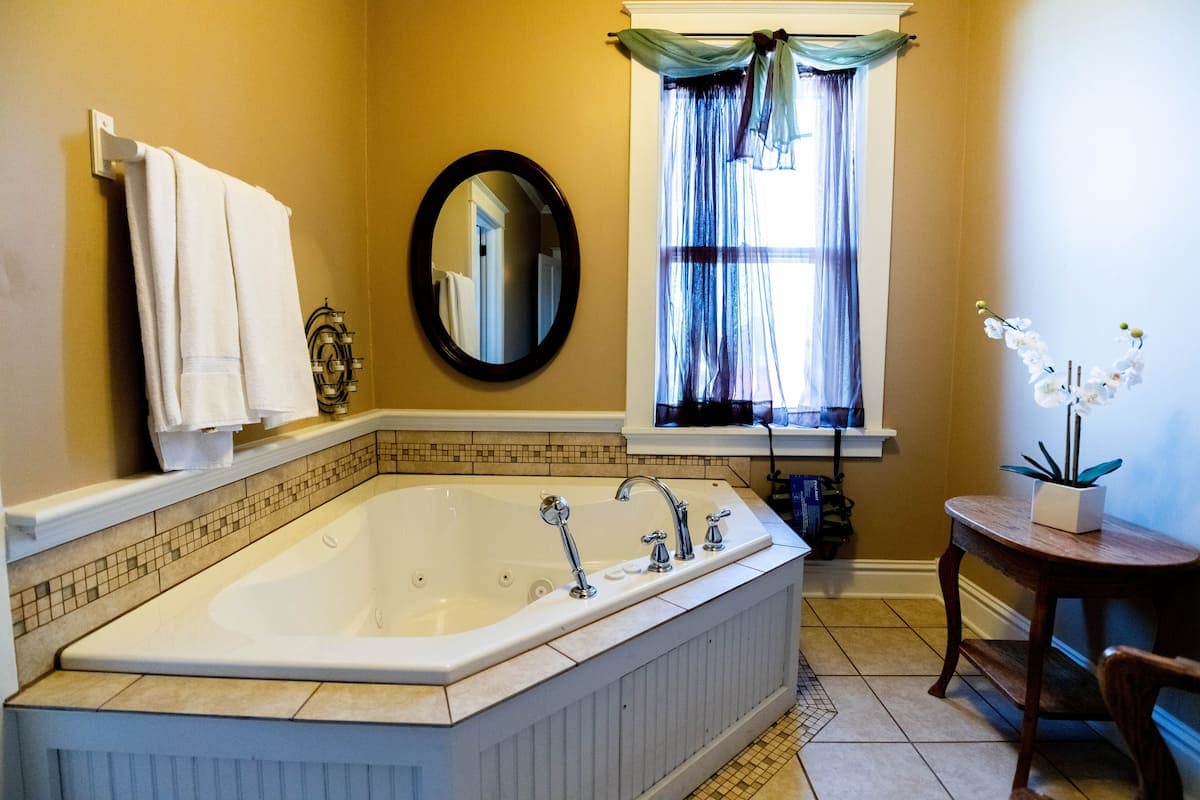 This elegant bathroom features a corner jetted tub, a round mirror, and a window with sheer dark blue drapes, complemented by light brown walls.