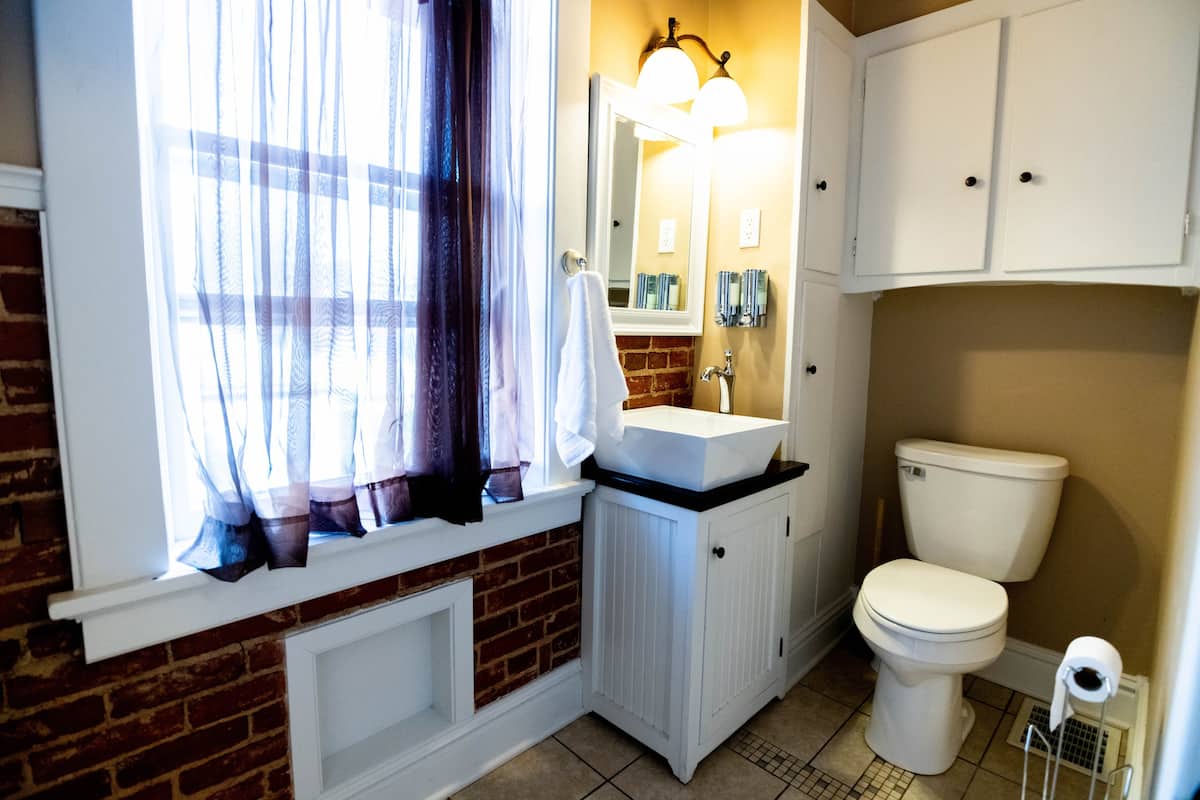 This cozy bathroom features exposed brick, a white vessel sink on a vanity, a toilet, built-in storage, and a large window with sheer curtains.