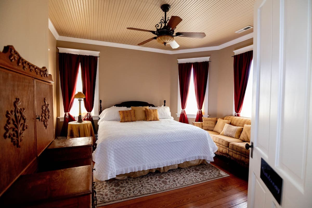 A classic bedroom featuring a large bed with white linens, a dark wood headboard, red curtains, a ceiling fan, and a cozy sofa.