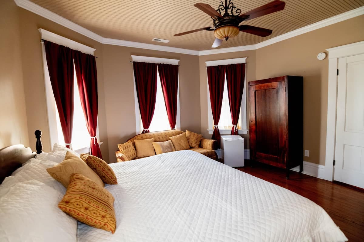 A spacious bedroom with a large bed, yellow accent pillows, a dark wood armoire, a sofa, a ceiling fan, and red drapes on multiple windows.