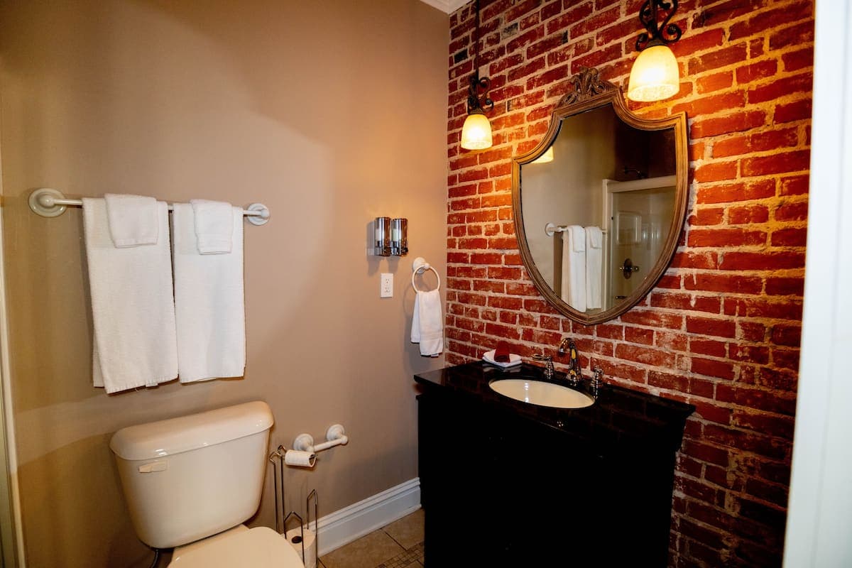 A stylish bathroom with an exposed brick accent wall, a dark vanity with a white sink, an ornate mirror, and a toilet.