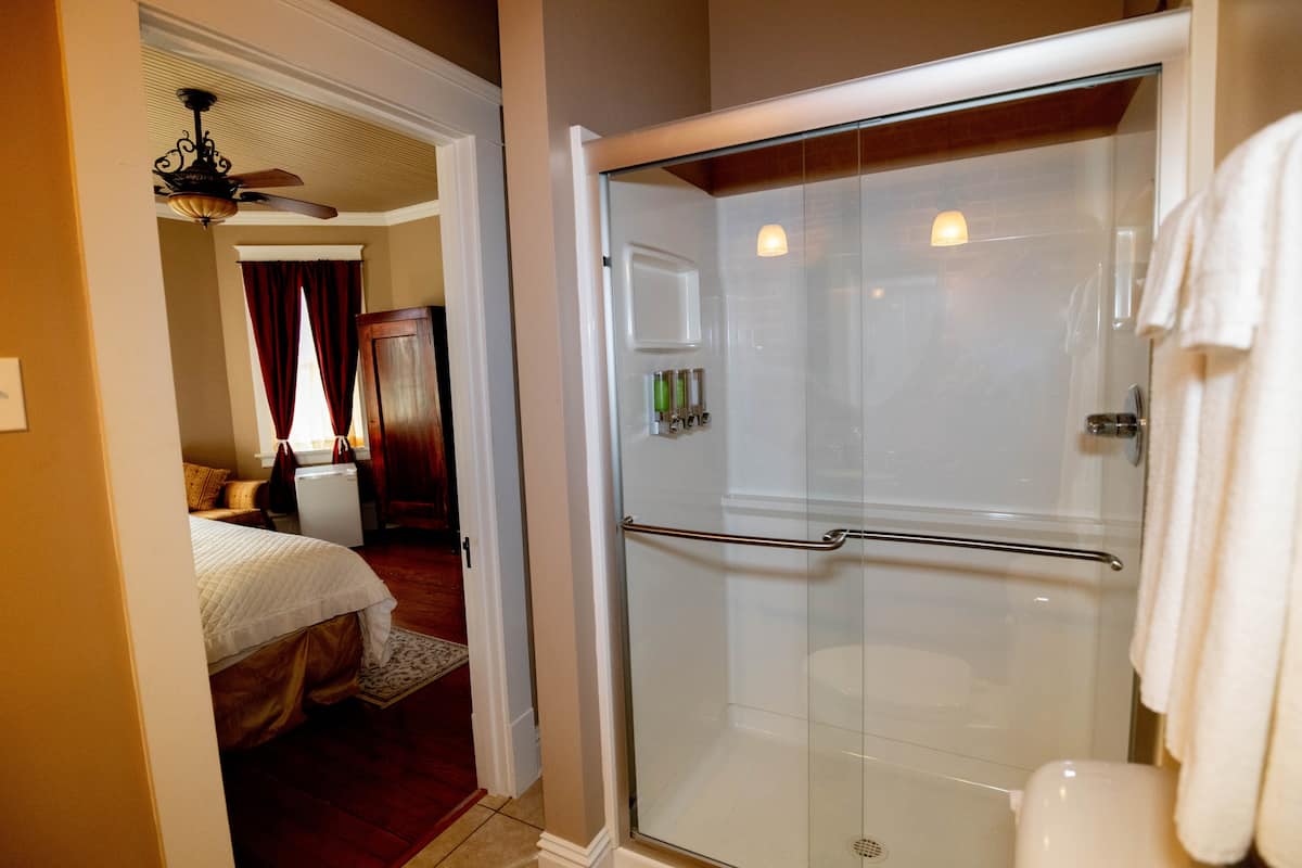 A bright bathroom with a glass-enclosed shower, a toilet, and white towels, with a view into an adjacent bedroom.