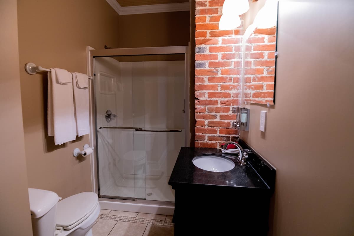 This functional bathroom features a glass-enclosed shower, a toilet, a vanity with a dark countertop and sink, and an exposed brick accent wall.
