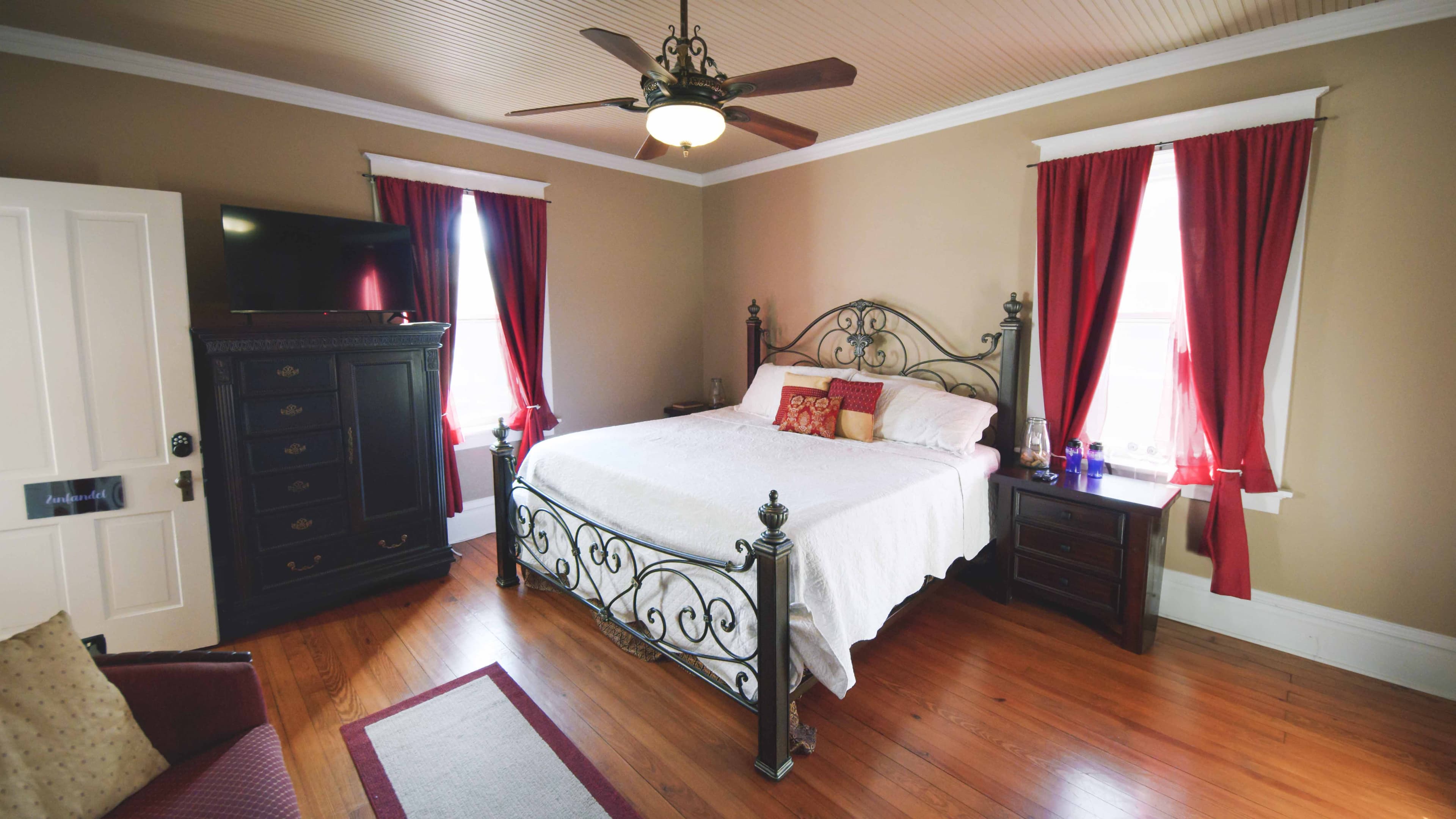 This cozy bedroom features a metal-frame bed with white bedding, dark wood furniture, rich red curtains, and hardwood floors.