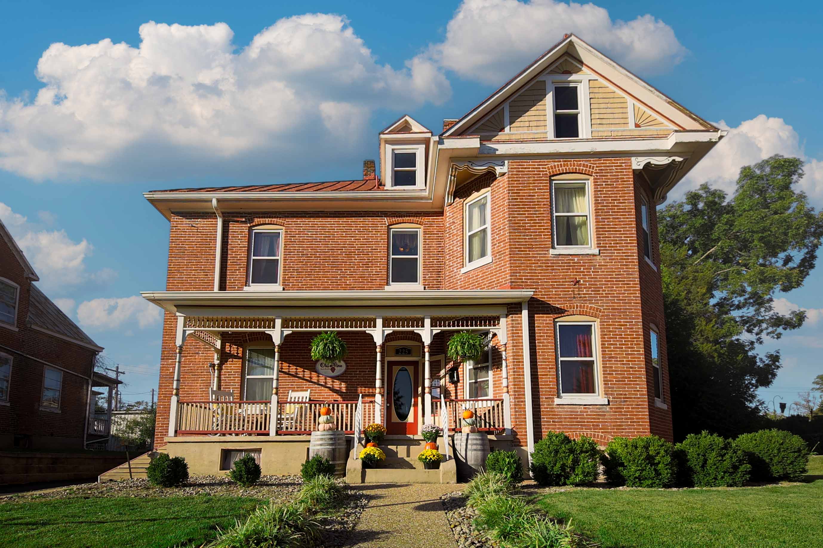 A grand red brick house with a prominent front porch, ornate white columns, and multiple windows, set against a blue sky with white clouds.