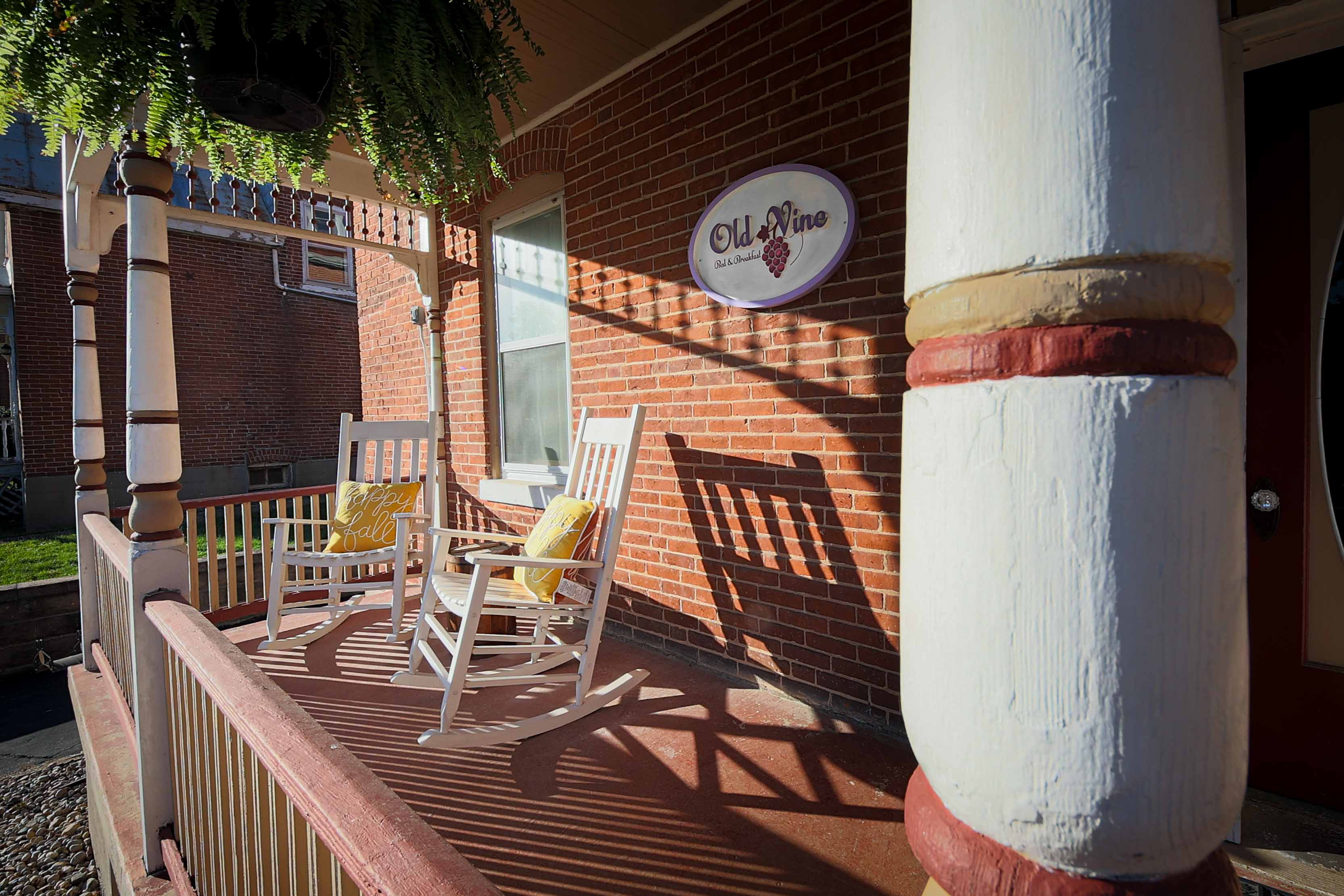 A inviting front porch with two white rocking chairs featuring yellow pillows, an "Old Vine" sign on the brick wall, and a hanging plant.