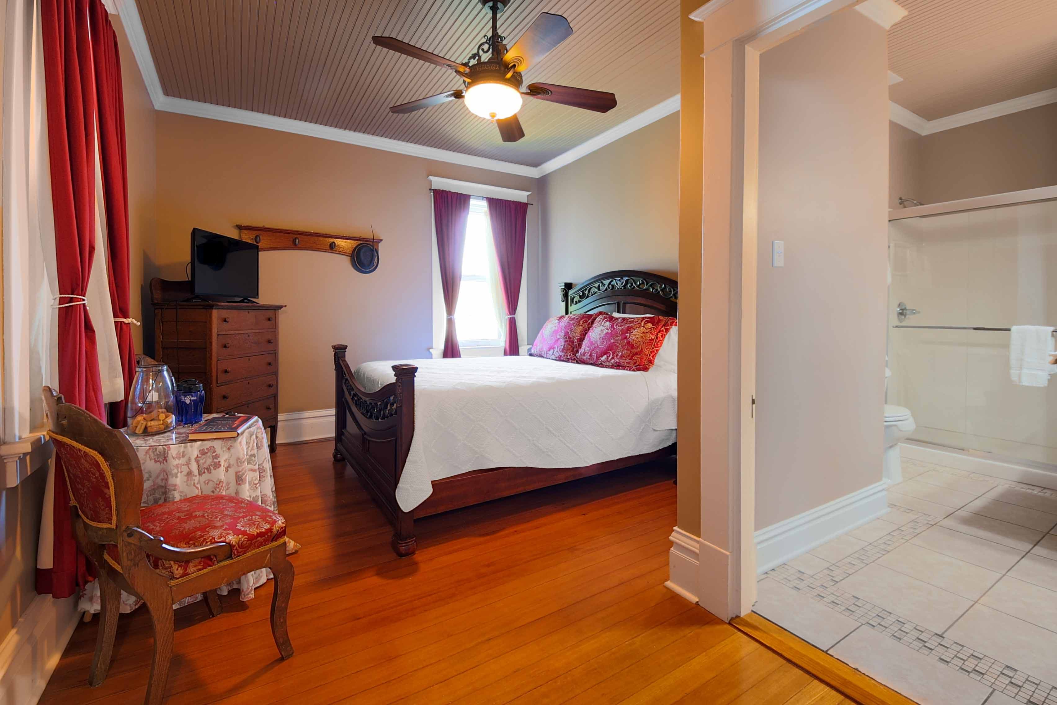 A charming bedroom featuring a bed with colorful pillows, hardwood floors, a ceiling fan, red drapes, and an adjacent bathroom visible.