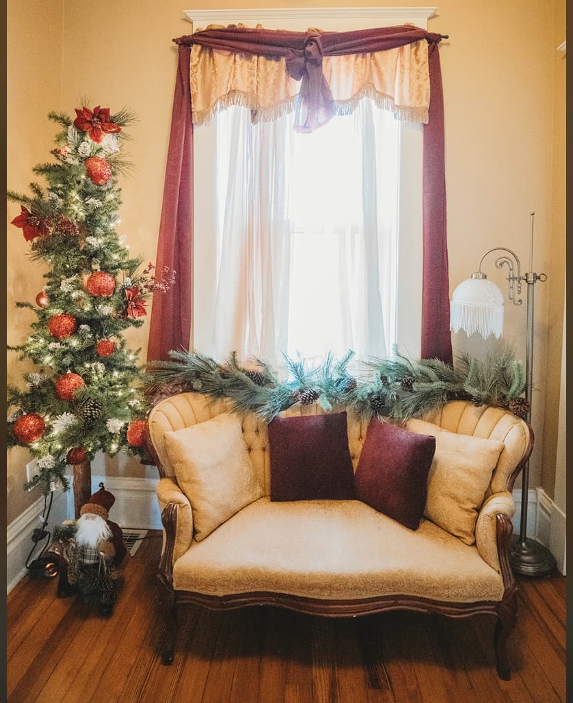 A cozy, decorated living space featuring a festive Christmas tree, a vintage cream-colored sofa with burgundy pillows, and soft natural light streaming through sheer curtains.