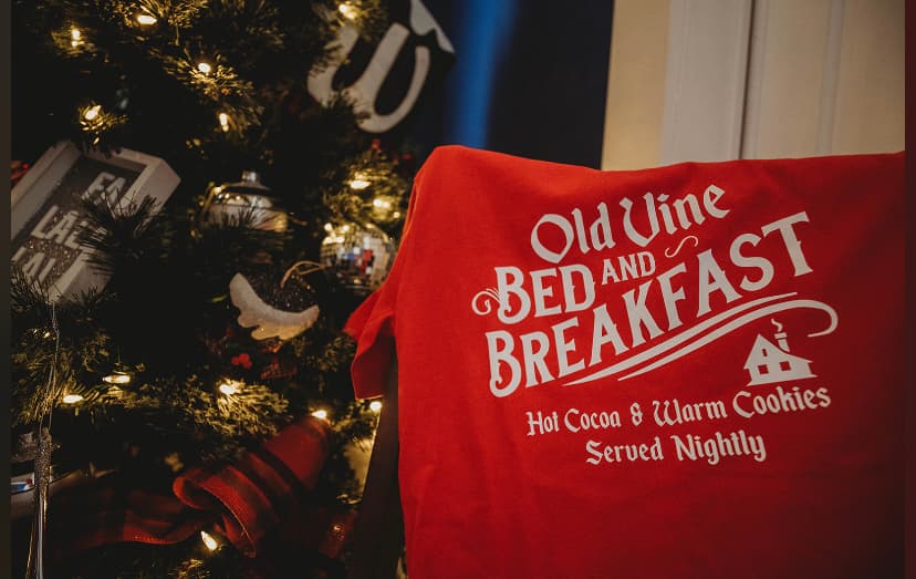 A red t-shirt with the text "Old Vine BED AND BREAKFAST - Hot Cocoa & Warm Cookies Served Nightly" displayed in front of a Christmas tree.