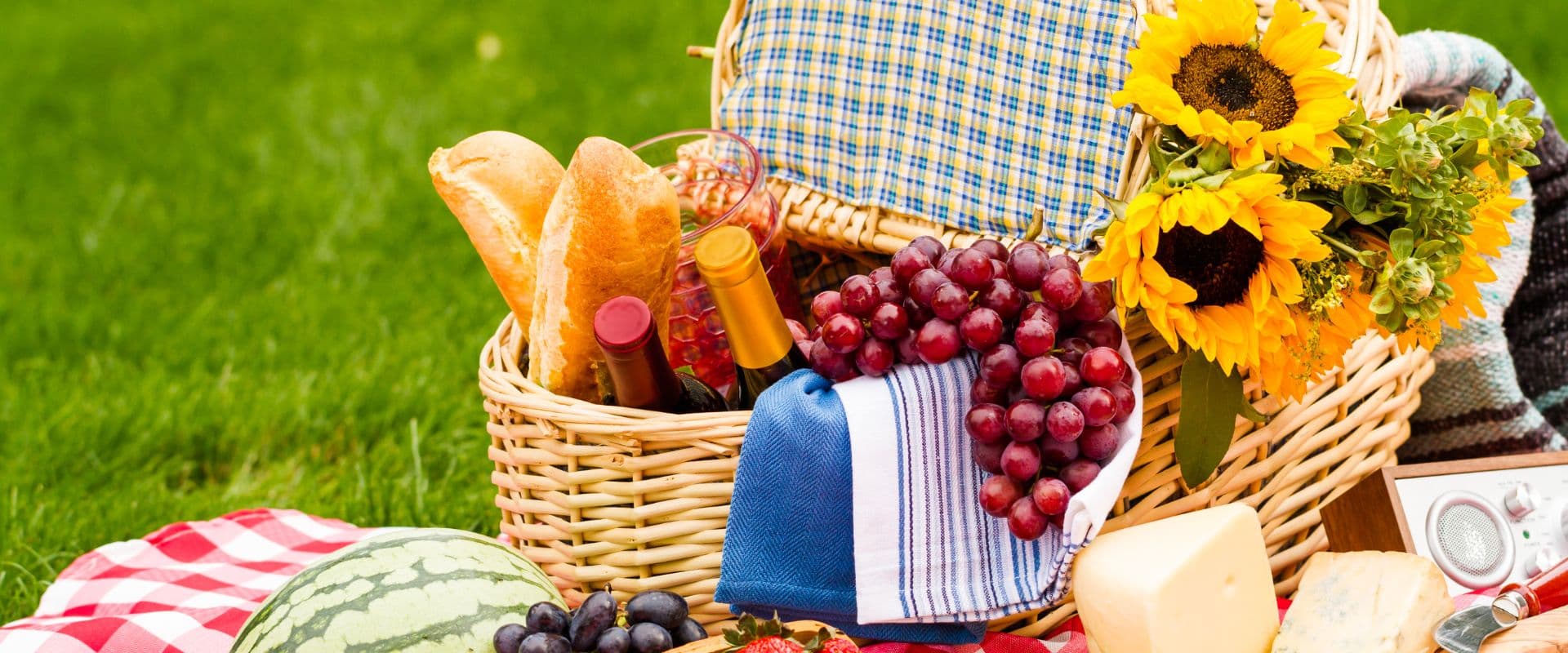A picnic basket filled with bread, wine, grapes, cheese, and sunflowers on a grassy background.
