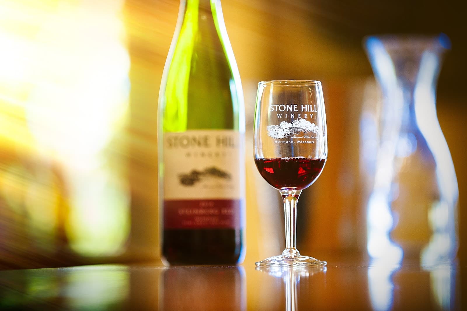 A bottle of Stone Hill wine next to a filled glass on a reflective surface.