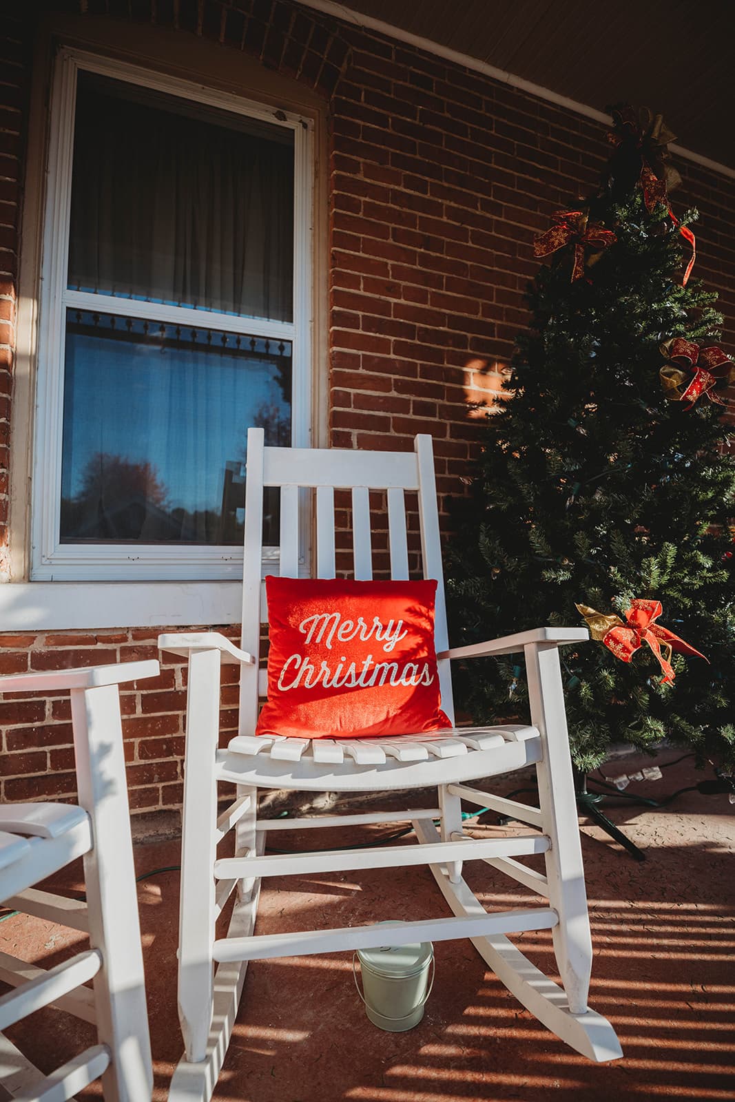 A white rocking chair with a red "Merry Christmas" pillow beside a decorated Christmas tree.