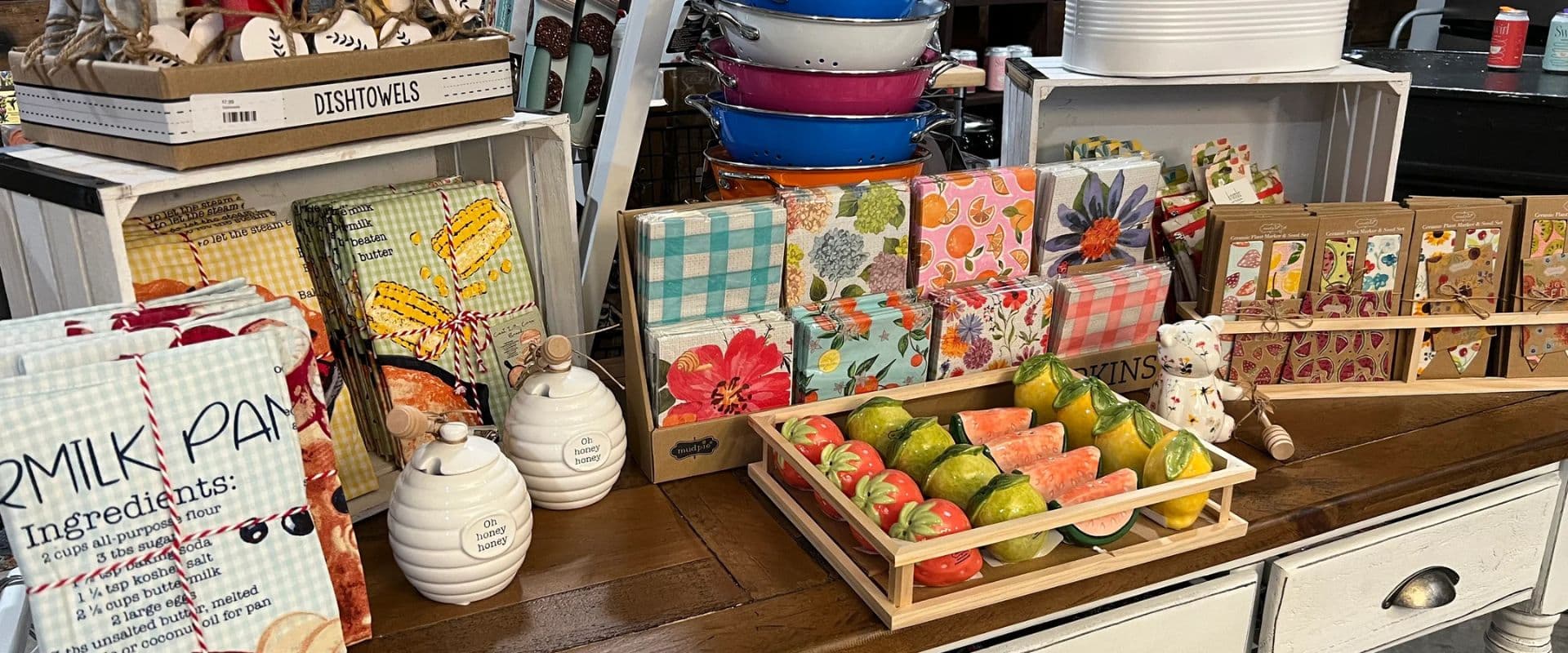 A display of colorful dish towels, napkins, decorative food items, and ceramic containers on a wooden table.