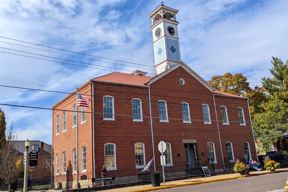 Historic red brick building with a clock tower and American flag, surrounded by trees and a sidewalk.