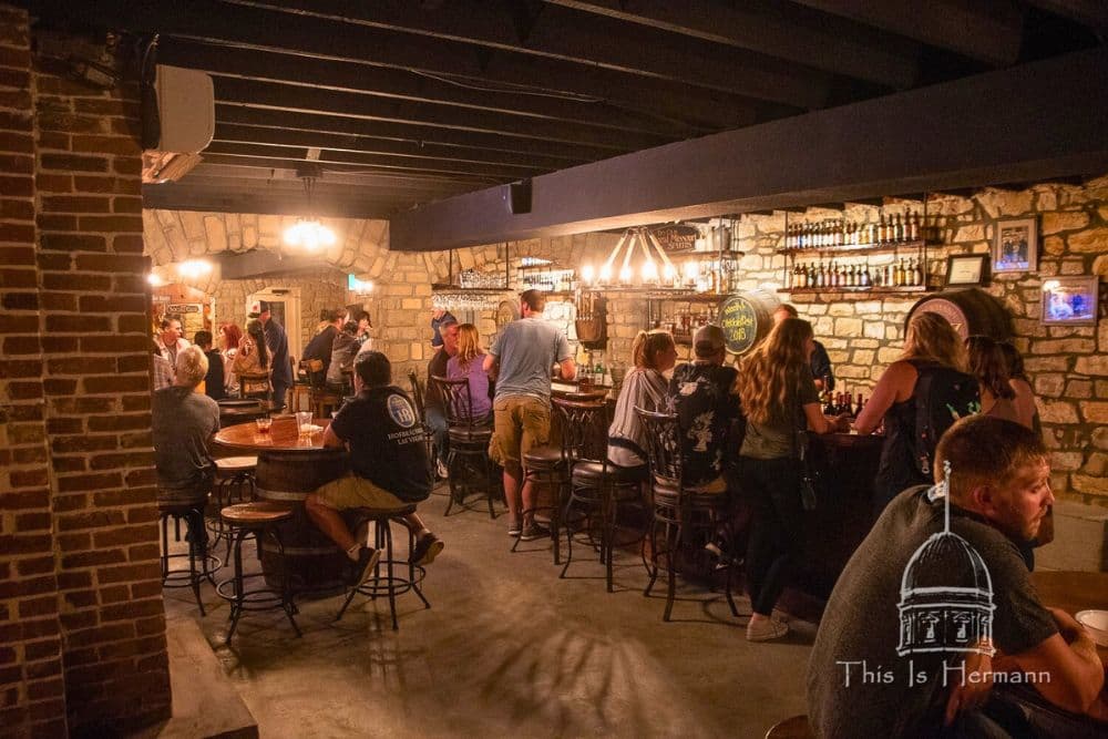 A bustling bar with patrons seated at tables and the bar, surrounded by stone walls and shelves of liquor.