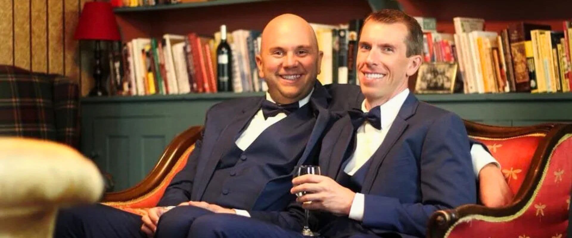 Two men in formal suits sit together on a couch, smiling and holding a glass.
