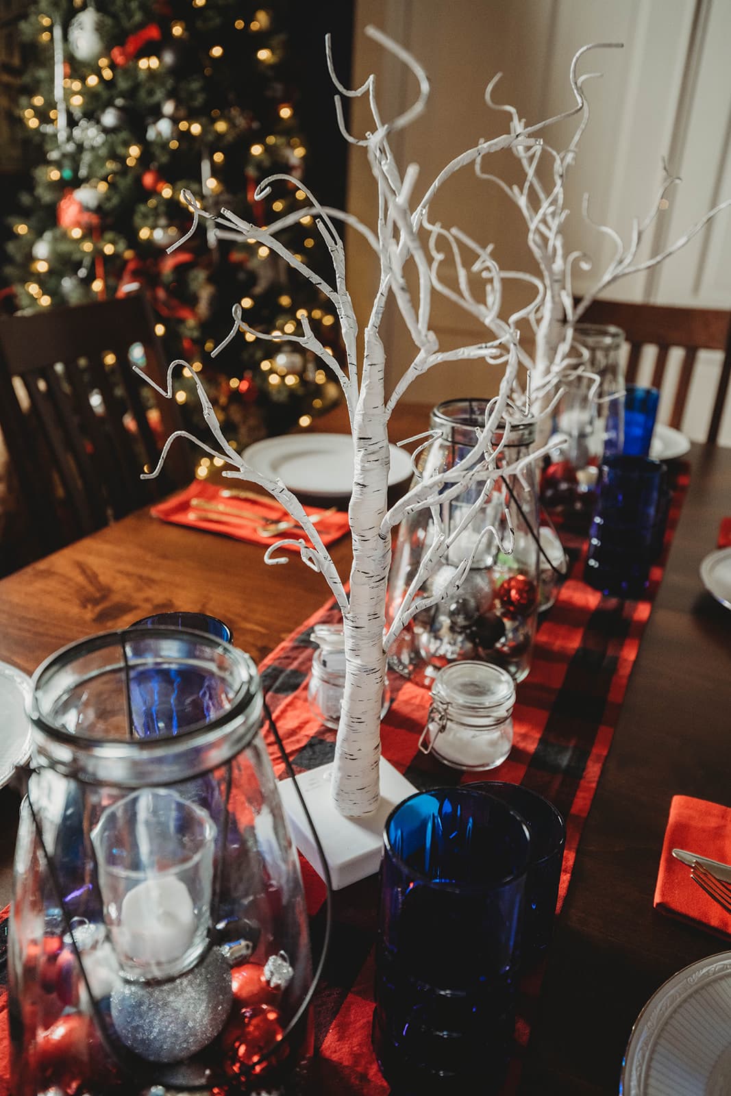 A festive dining table decorated with white branched trees, glass jars, blue glasses, and a red and black checkered table runner, with a Christmas tree in the background.